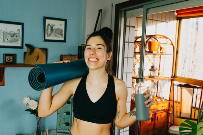 Woman carrying a yoga mat and water bottle, smiling at the camera