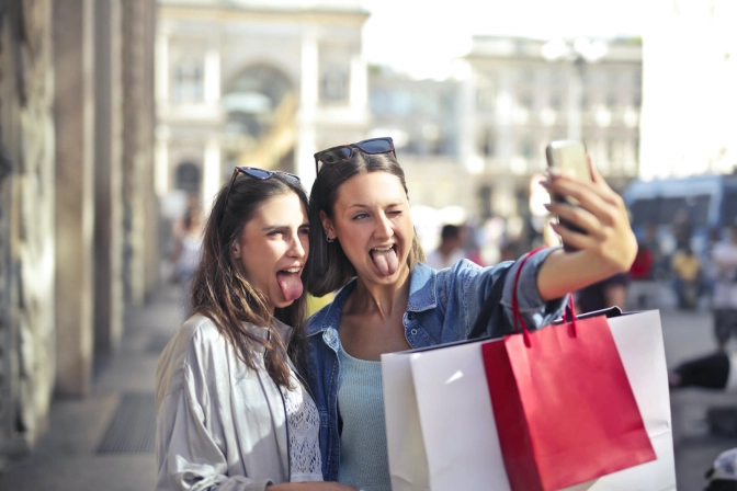 A woman stretching her arm out to take a selfie with her friend with shopping bags around her arms.&nbsp;