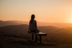 sillhouette of woman sitting on bench while watching the sunset on tall mountains