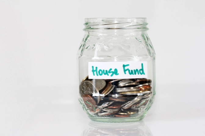 Transparent glass jar labeled "House Fund", half-filled with coins