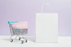 a pink sale tag placed inside a shopping cart figurine next to a white shopping bag on a white table
