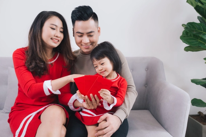 Family of three sitting on a grey couch holding three red packet envelopes&nbsp;