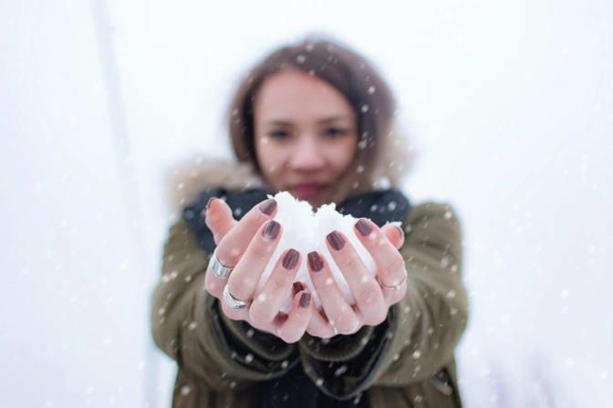 Brunette woman with maroon nail polish holding a pile of snow in the winter